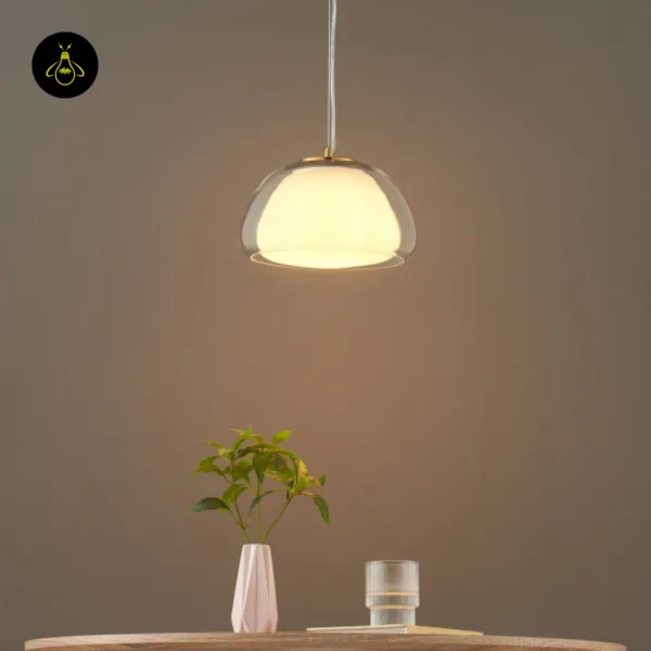 Lomelia double glass pendant light lit above a wooden table with a white vase and fluted glass, front view