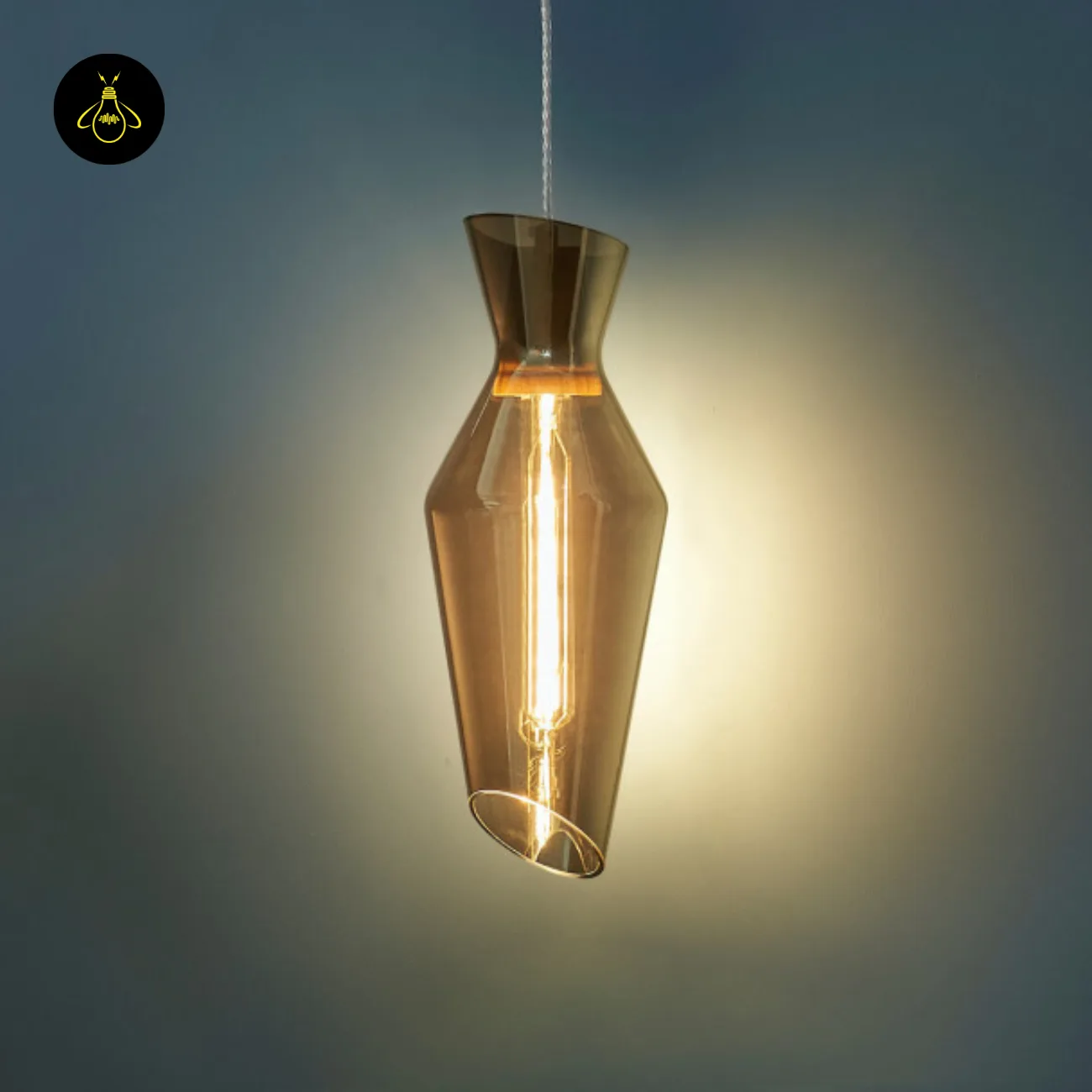 Faceted Smoked Glass Pendant Light – Loft Drop Form, 40 cm 6 Faceted smoked glass pendant light with a slant-cut lower rim and warm filament glow against a blue-grey wall
