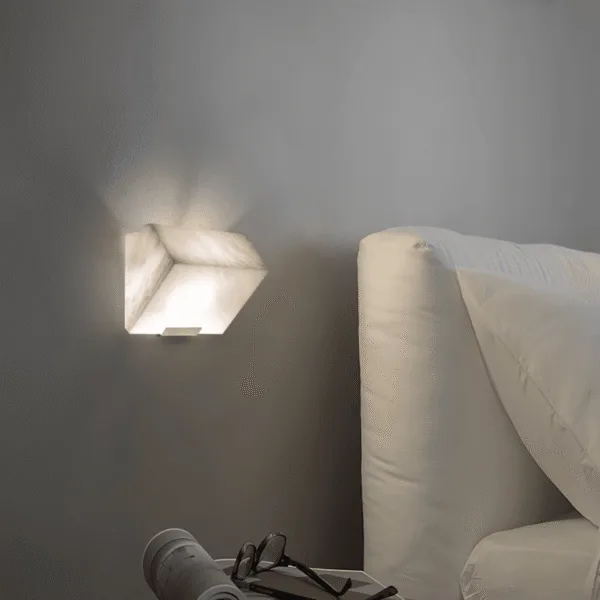 Alabaster stone wall light lit beside a white upholstered bed, soft bedside light over a side table with glasses