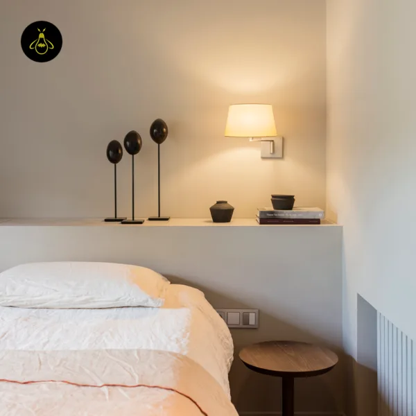 Swing arm wall lamp lit on a bedroom wall above a ledge, beside a bed and small round side table