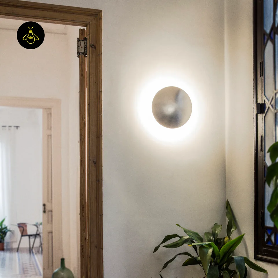 Round LED Wall Light – Satin Nickel Eclipse Disc, 24 cm Warm White Halo 5 LED wall light glowing on a white hallway wall beside a wooden door frame and a potted plant