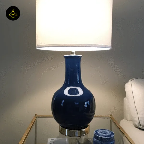 Royal blue ceramic table lamp lit with a warm glow on a glass side table