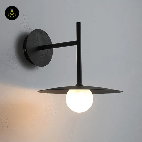 Close-up of the black wall lamp glowing warm on a grey wall, frosted globe lit under the disc