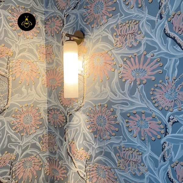 Gold-lustre borosilicate glass wall light with frosted opal cylinder lit against a blue and pink floral patterned wallpaper