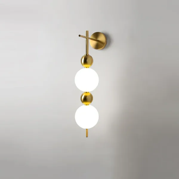Bedside wall light lit on a grey wall, its opal borosilicate glass globes glowing between brushed-brass beads