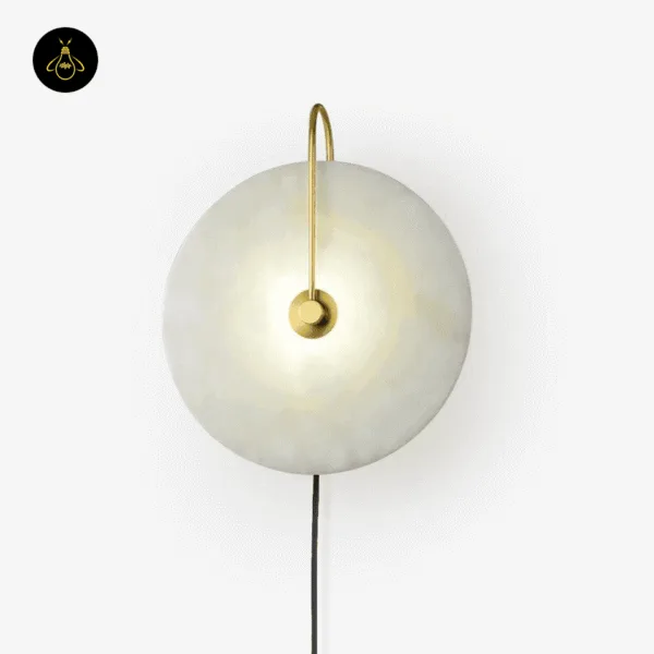 Gold plug in wall lamp lit on a white wall, black cord running down from the alabaster disc