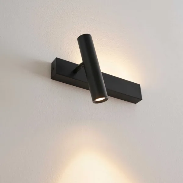 Matte black wall reading lamp with the reading head angled down and the backplate glowing up the wall