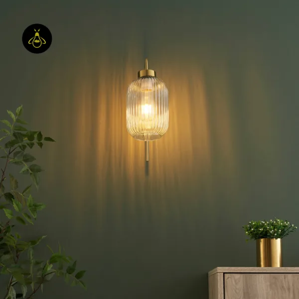 Front view of a fluted glass wall light glowing on a dark green wall, gold rod below the shade