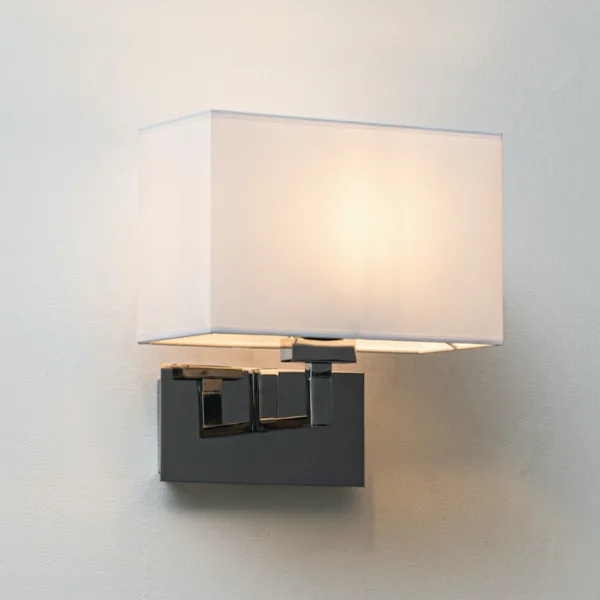 Wall lamp with shade glowing warm: white fabric shade over a polished chrome stainless steel arm and backplate