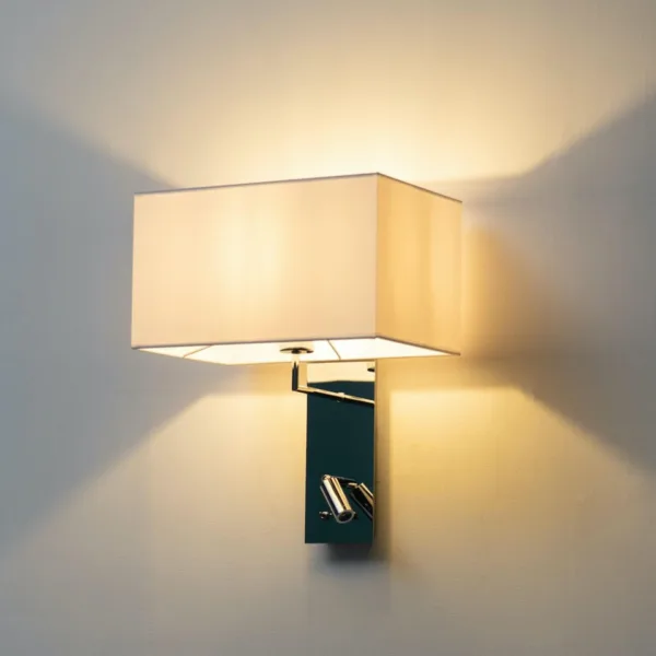 Wall mounted bedside light glowing warm through its white fabric shade above a chrome backplate