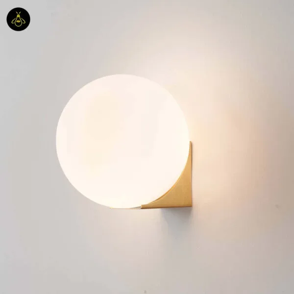 Globe wall light lit, white opal ball on a folded brushed-gold base, three-quarter view