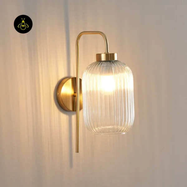 Close-up of a lit fluted glass wall light, clear ribbed glass barrel on a brushed gold stainless steel arm