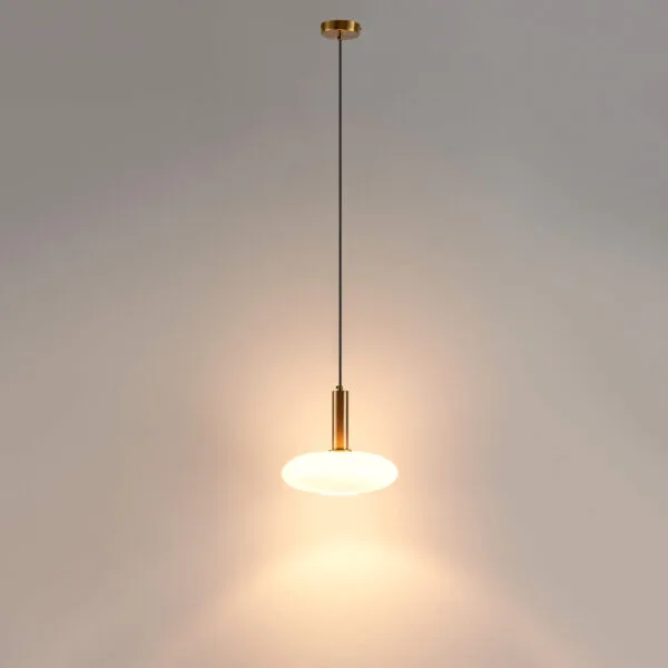 Opaline glass pendant light glowing on a long black cable below its round brushed gold canopy, against a beige wall