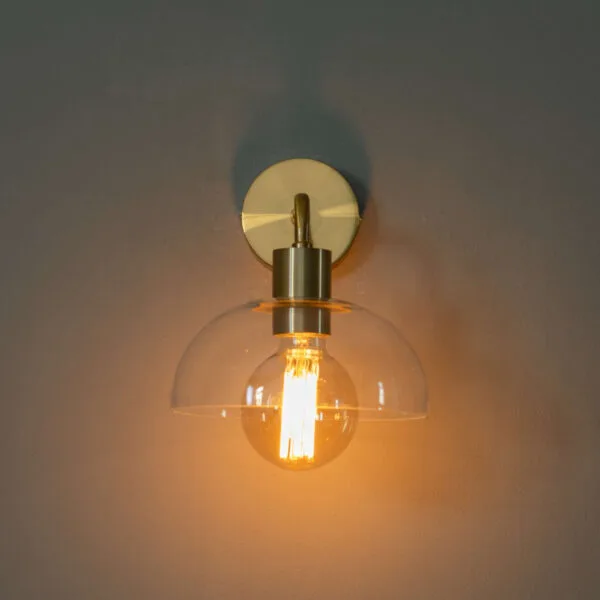 Front view of the dome wall light lit on a grey wall, round antique brass finish backplate above a clear glass dome
