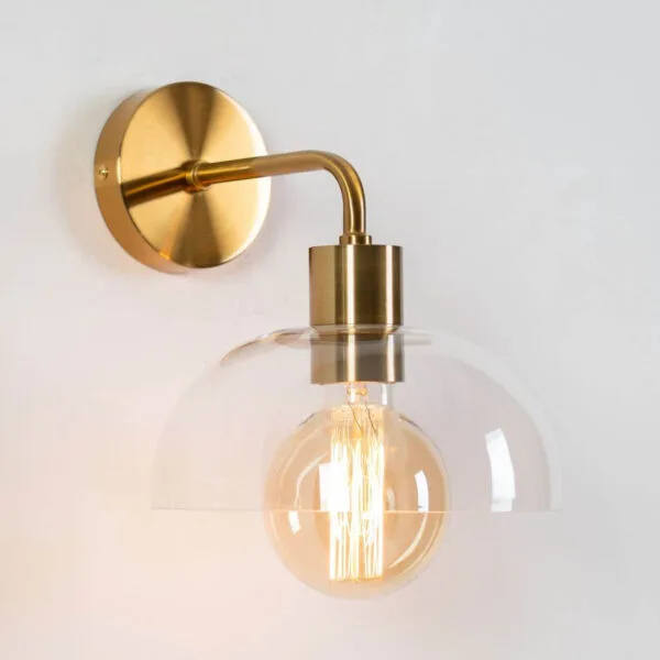 Dome wall light at a three-quarter angle: clear borosilicate glass dome, curved arm and round backplate in antique brass finish