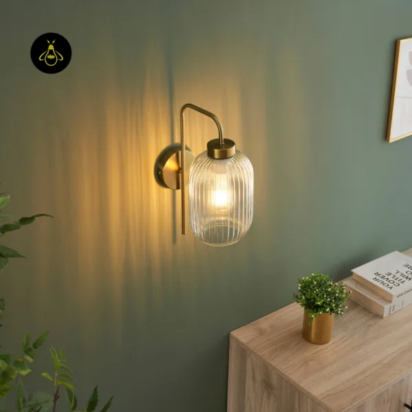 Fluted glass wall light lit at an angle, clear ribbed glass shade on a brushed gold arm, wooden sideboard with books below