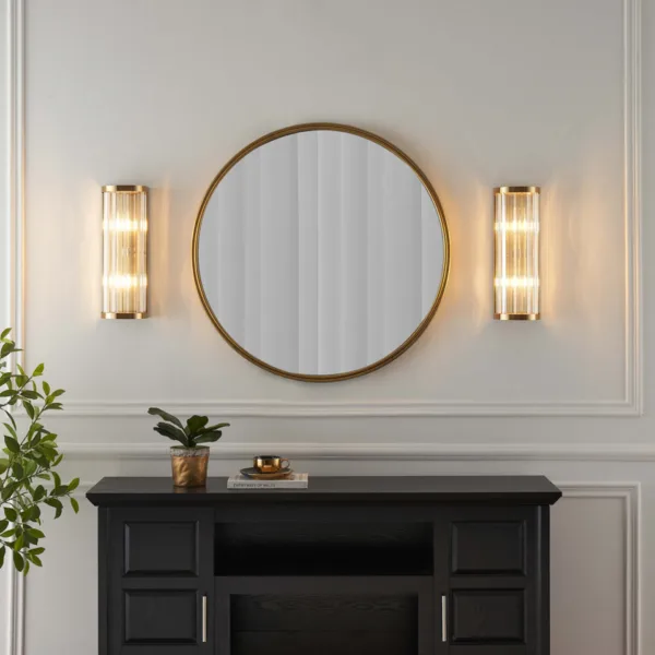 Wide view of two ribbed glass wall lights glowing beside a round gold mirror over a black console
