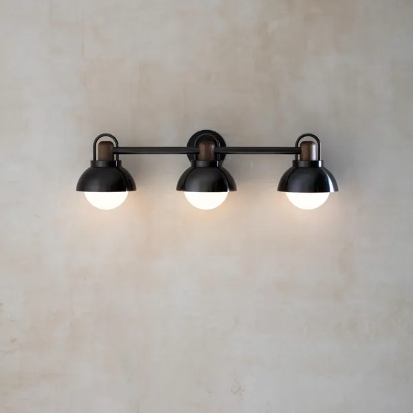 Vanity wall light with three lit opal borosilicate glass globes under black steel domes on a plaster wall, front view