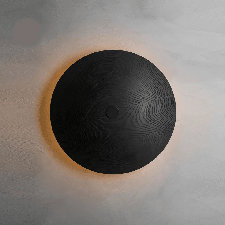 Wooden wall light from the front: round 51 cm black wood disc with a warm halo glowing on a grey plaster wall