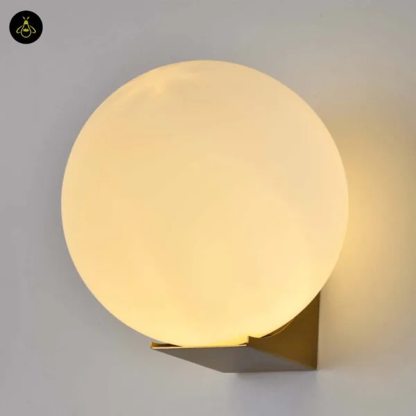 Globe wall light lit, warm glow filling the opal borosilicate glass ball above the brushed-gold base
