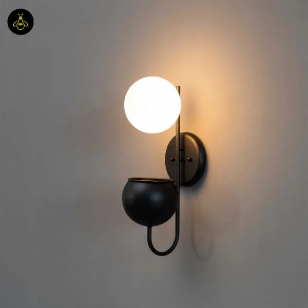 Plant pot wall light seen from the left, warm glow on a plain wall above the matte black planter cup
