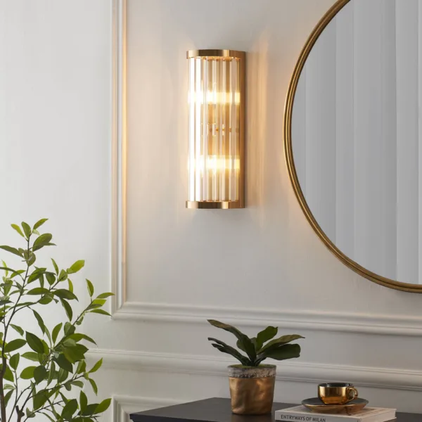 Gold sconce with clear glass rods glowing on a panelled wall beside a round gold mirror, ribbed glass wall light above a console