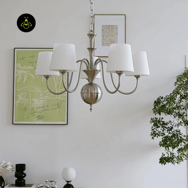 White chandelier unlit in a dining room, with framed prints on the wall and a green plant