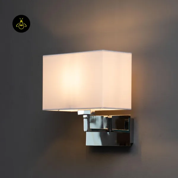 Wall lamp with shade lit front-on, the white fabric glowing warm over a polished chrome backplate
