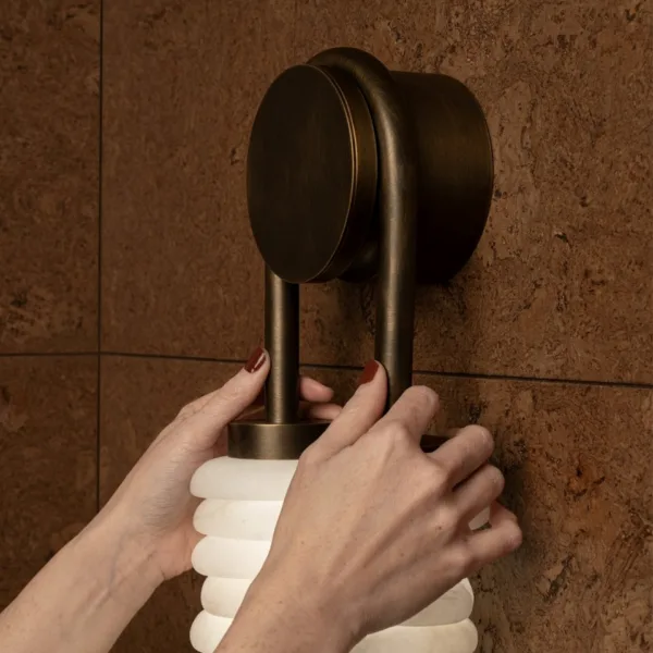 Hands fitting a column of the stacked alabaster wall light, showing the round backplate and U-arm in aged-brass finish on 304 stainless steel