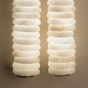 Designer wall light front view with glowing stacked alabaster cylinders and warm brass round backplate