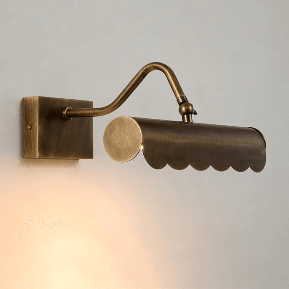 Brass mirror light lit warm on a plain wall, scalloped shade on a swan-neck arm