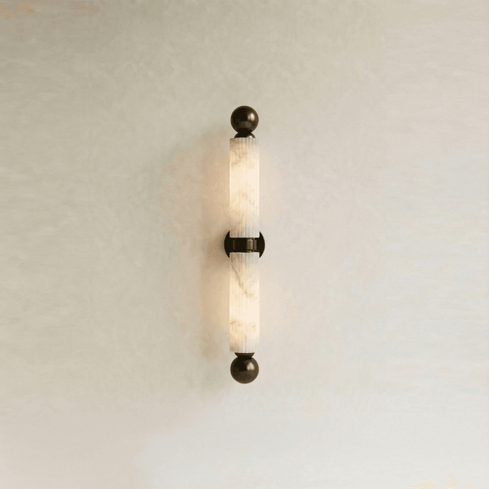 Fluted alabaster wall light lit warm, twin natural stone tubes with antique bronze spheres