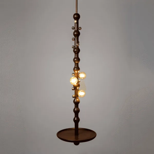 Teak wooden hanging lamp lit against a grey wall, three amber borosilicate glass globes glowing warm along the turned teak stem