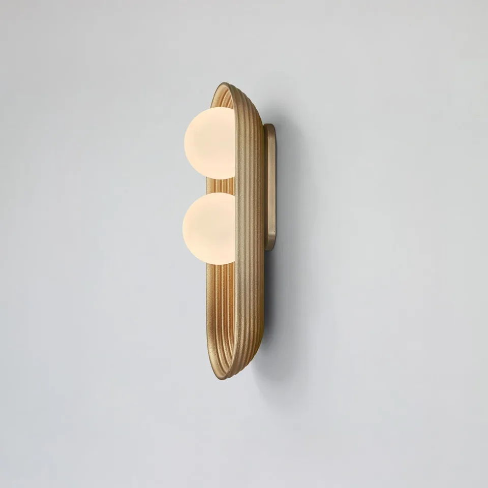 Twin oval wall light in matt gold, reeded wood capsule backplate with two opal glass balls, on a grey wall