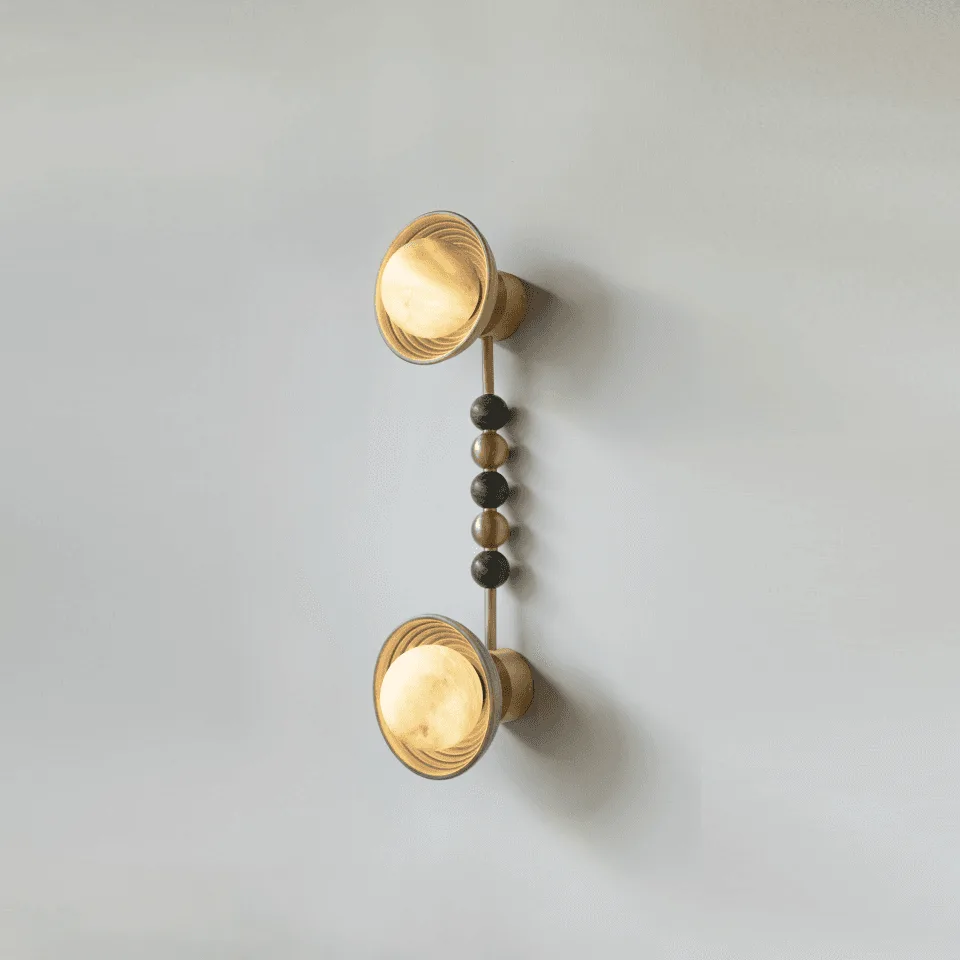 Twin wall sconce with two reeded matt-gold wood bowls and opal glass balls lit, joined by a stem of gold-metal and green stone beads, on a plain wall