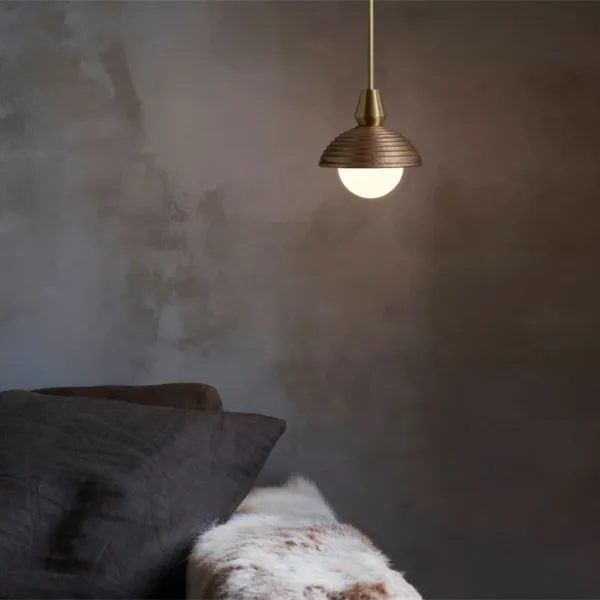 Wood pendant light glowing warm beside a bed, opal ball under the reeded dome, grey pillows and a fur throw below