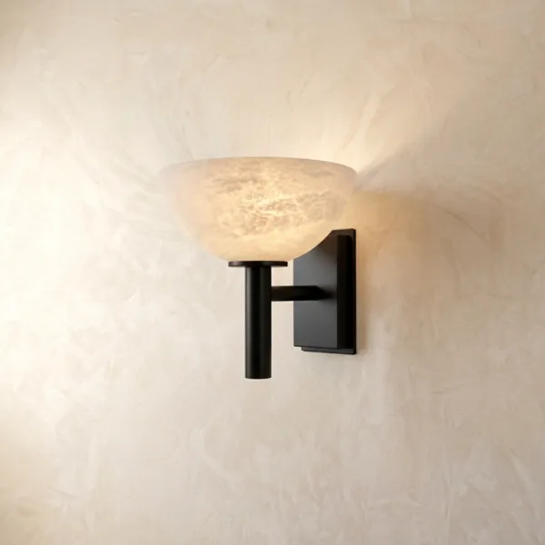Alabaster bowl wall light lit, warm light washing up a textured plaster wall from the stone bowl on a matte black stem