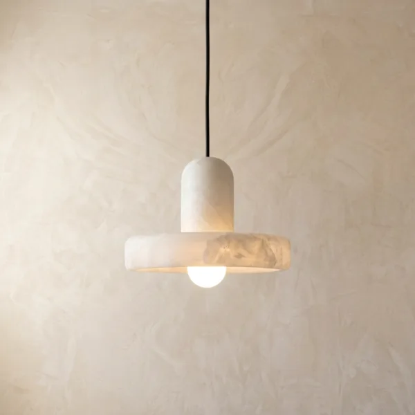 Alabaster pendant lamp lit, its white stone disc and rounded cap glowing over a globe bulb against a plaster wall