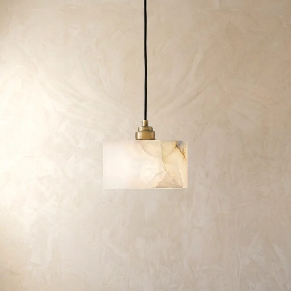 Natural alabaster drum pendant light with a brass socket cup glowing against a plaster wall, showing soft white veining