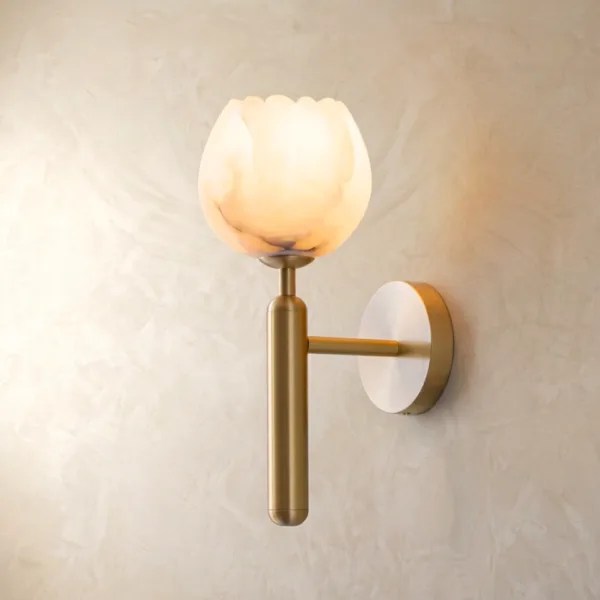 Tulip wall light in natural alabaster glowing warm on a brass-finish stainless steel stem against a plaster wall