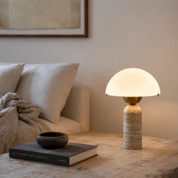 Travertine table lamp with a glowing opal glass dome on a bedside table beside cushions