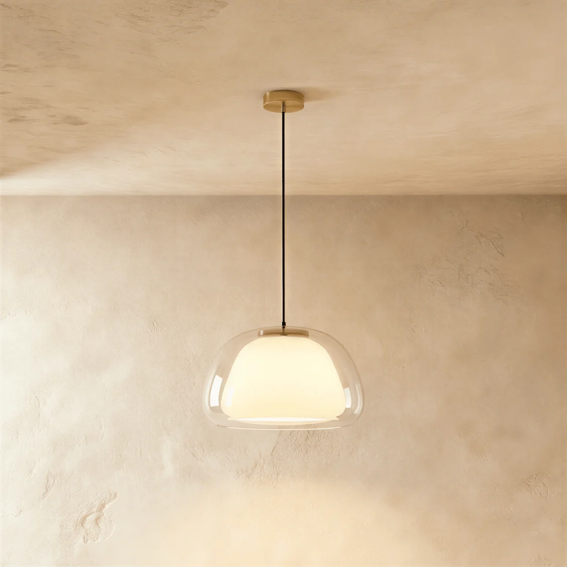 Double glass pendant light with a clear outer shade and glowing opal inner shade on a gold canopy, against a plaster wall