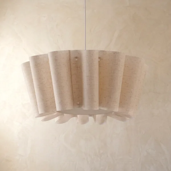 Draped fabric pendant light with a natural-weave shade of long soft folds and a ruffled hem against a plaster wall