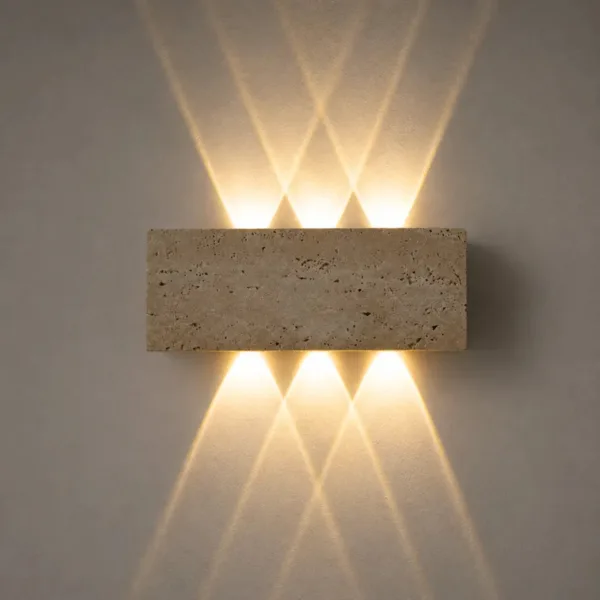 Outdoor up and down light in honed travertine on a grey wall, its pitted stone face lit warm