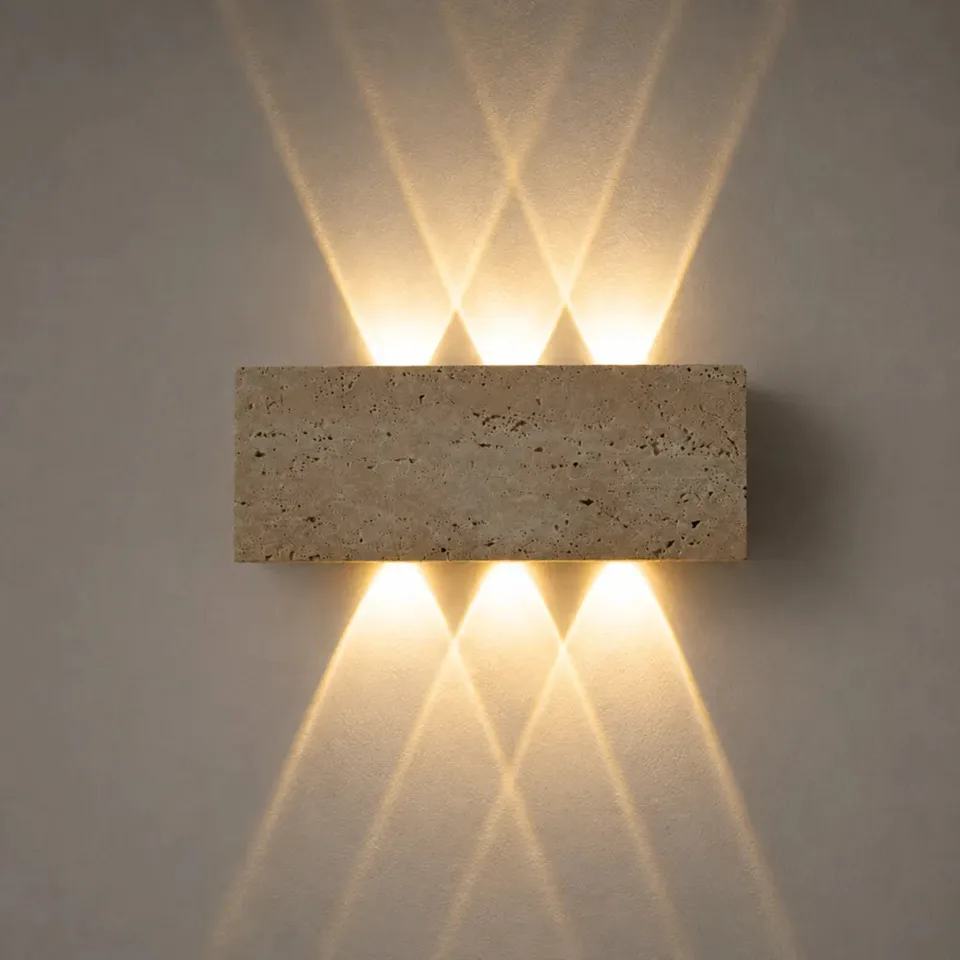Outdoor up and down light in honed travertine on a grey wall, its pitted stone face lit warm