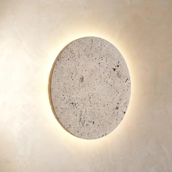 Travertine disc wall light glowing with a warm halo on a plaster wall, its pitted cream stone face unlit