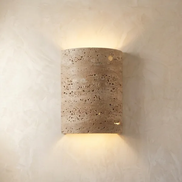 Half cylinder wall sconce in travertine glowing up and down on a cream plaster wall, the pitted stone face unlit