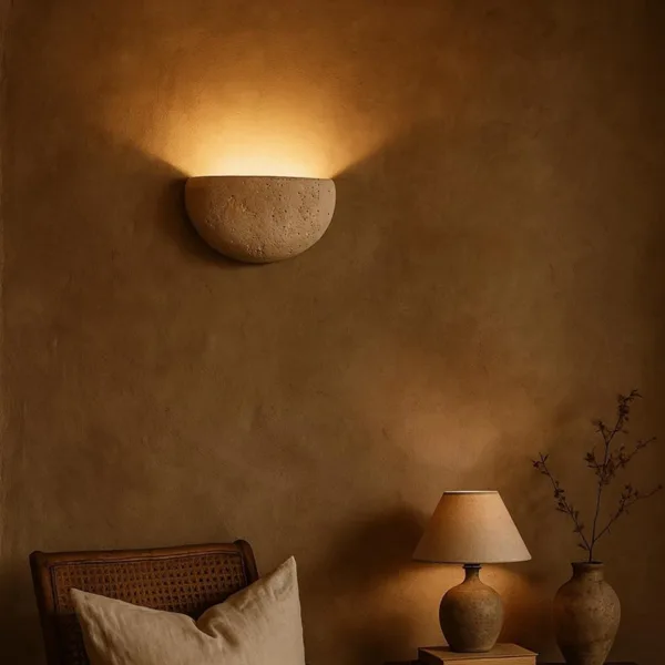 Travertine moon wall light on an ochre plaster wall above a rattan chair and a stone table lamp in a sitting room