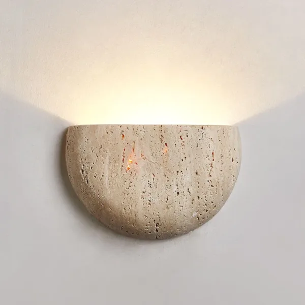 Travertine moon wall light on a white wall, warm uplight above and small points of light glowing through the stone pits